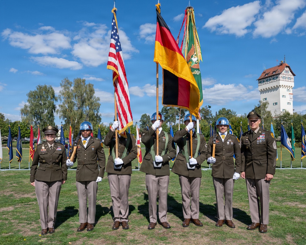 A formal military color guard ceremony featuring U.S. Army personnel in dress uniforms holding the American flag and the German national flag, along with additional unit guidons. The ceremony appears to take place on a parade ground in what appears to be a German town, evidenced by the traditional half-timbered tower building visible in the background. Personnel are arranged in a formal line, with flag bearers wearing blue ceremonial helmets.