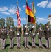 A formal military color guard ceremony featuring U.S. Army personnel in dress uniforms holding the American flag and the German national flag, along with additional unit guidons. The ceremony appears to take place on a parade ground in what appears to be a German town, evidenced by the traditional half-timbered tower building visible in the background. Personnel are arranged in a formal line, with flag bearers wearing blue ceremonial helmets.