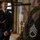 Two military personnel in dress uniforms are engaged in conversation indoors. The foreground figure wears a U.S. Army olive dress uniform with a '1st Infantry Division' shoulder patch and Staff Sergeant chevrons, and is holding a brass bugle. The background figure wears a dark navy/black ceremonial uniform with gold aiguillette, likely from a European military.