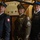 Three military personnel pose together indoors, two wearing Polish Armed Forces dress uniforms with blue caps and white eagle patches, and one wearing a U.S. Army dress uniform with olive green coat, service ribbons, and Staff Sergeant rank insignia. The setting appears to be a historic or ceremonial interior with wooden beam architecture and a framed document or certificate visible on the wall behind them. The occasion appears to be a formal military or diplomatic event.