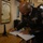 A U.S. Army senior non-commissioned officer in dress uniform is signing what appears to be an official guest book or register at a formal location. The soldier displays multiple service ribbons, a unit patch identified as the 1st Infantry Division, and sergeant first class or master sergeant rank chevrons. A framed Latin document titled 'PRAECEPTUM' is visible on the wall behind him.