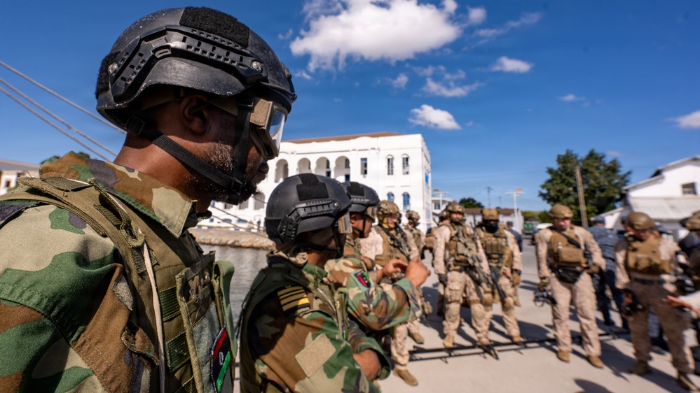 The image shows a group of armed military personnel in tactical gear assembled in an outdoor urban area. Two distinct groups appear to be present: soldiers wearing woodland camouflage with black tactical helmets, and others in desert/tan camouflage uniforms. The setting appears to be a courtyard or plaza near a white colonial-style building.