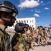 The image shows a group of armed military personnel in tactical gear assembled in an outdoor urban area. Two distinct groups appear to be present: soldiers wearing woodland camouflage with black tactical helmets, and others in desert/tan camouflage uniforms. The setting appears to be a courtyard or plaza near a white colonial-style building.