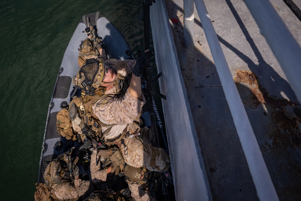 An overhead aerial photograph shows multiple armed military personnel in full tactical gear aboard a rigid inflatable boat (RIB) positioned alongside a dock or pier structure. One soldier in desert camouflage uniform is reaching upward toward the dock edge, suggesting a boarding or infiltration exercise. The scene appears to depict a maritime special operations training or tactical exercise.