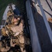An overhead aerial photograph shows multiple armed military personnel in full tactical gear aboard a rigid inflatable boat (RIB) positioned alongside a dock or pier structure. One soldier in desert camouflage uniform is reaching upward toward the dock edge, suggesting a boarding or infiltration exercise. The scene appears to depict a maritime special operations training or tactical exercise.