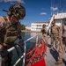 Military personnel, appearing to be U.S. Marines and possibly allied forces, are conducting a maritime boarding or vessel inspection exercise at a port facility. One heavily equipped operator in full tactical gear is climbing over a ship's railing using a red boarding ladder, while several other personnel observe from the dock. The setting appears to be a tropical or Mediterranean-style port with white colonial-style buildings visible in the background.