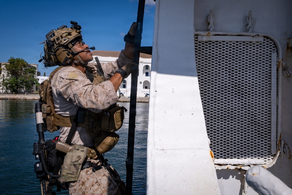 A heavily equipped military service member in MARPAT (Marine pattern) camouflage uniform is climbing or scaling the side of a white vessel near a waterfront. The individual is wearing full tactical gear including a combat helmet with night vision mount, communications headset, body armor, and multiple pouches. A white building and blue water are visible in the background.