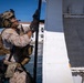 A heavily equipped military service member in MARPAT (Marine pattern) camouflage uniform is climbing or scaling the side of a white vessel near a waterfront. The individual is wearing full tactical gear including a combat helmet with night vision mount, communications headset, body armor, and multiple pouches. A white building and blue water are visible in the background.
