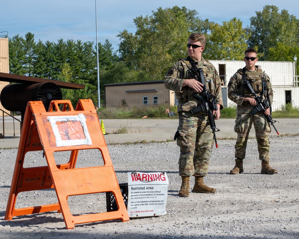 Two armed military personnel in camouflage uniforms stand guard at what appears to be a restricted area checkpoint on a gravel surface. They are positioned near an orange warning sign stand displaying a 'WARNING - Restricted Area' notice. The setting appears to be a military installation or training facility with industrial buildings and trees visible in the background.