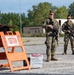 Two armed military personnel in camouflage uniforms stand guard at what appears to be a restricted area checkpoint on a gravel surface. They are positioned near an orange warning sign stand displaying a 'WARNING - Restricted Area' notice. The setting appears to be a military installation or training facility with industrial buildings and trees visible in the background.