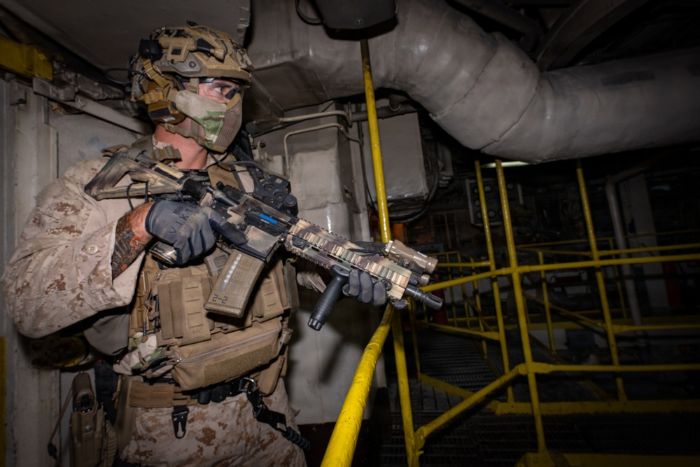 A heavily equipped military operator in desert MARPAT camouflage is conducting what appears to be a maritime vessel clearance or Visit, Board, Search, and Seizure (VBSS) operation in an industrial interior space. The operator is in a tactical ready position holding a camouflage-patterned assault rifle with multiple accessories. The environment features yellow safety railings, large pipes, and metal staircases consistent with a ship's interior or industrial facility.