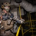 A heavily equipped military operator in desert MARPAT camouflage is conducting what appears to be a maritime vessel clearance or Visit, Board, Search, and Seizure (VBSS) operation in an industrial interior space. The operator is in a tactical ready position holding a camouflage-patterned assault rifle with multiple accessories. The environment features yellow safety railings, large pipes, and metal staircases consistent with a ship's interior or industrial facility.