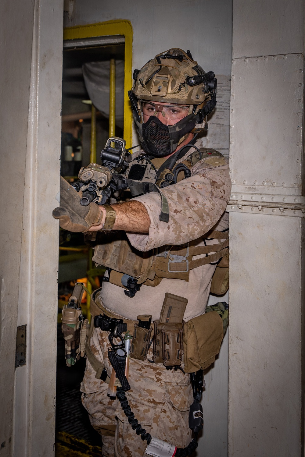 A heavily equipped military or special operations personnel is conducting what appears to be a maritime vessel boarding or clearance operation. The operator is positioned in a doorway/hatchway of what appears to be a ship's interior, holding a weapon at the ready. The individual is wearing full tactical kit including desert camouflage consistent with USMC MARPAT pattern.