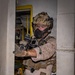 A heavily equipped military or special operations personnel is conducting what appears to be a maritime vessel boarding or clearance operation. The operator is positioned in a doorway/hatchway of what appears to be a ship's interior, holding a weapon at the ready. The individual is wearing full tactical kit including desert camouflage consistent with USMC MARPAT pattern.