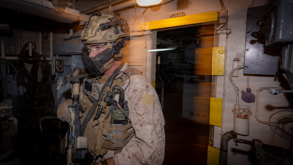 A heavily equipped military operator, wearing MARPAT desert camouflage consistent with U.S. Marine Corps uniforms, stands in a dimly lit interior corridor of what appears to be a naval vessel or ship. The operator is fully kitted with tactical gear including a helmet-mounted night vision device, face mask, body armor, radio equipment, and a slung weapon. The environment suggests a maritime boarding or ship-clearing operation.