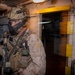 A heavily equipped military operator, wearing MARPAT desert camouflage consistent with U.S. Marine Corps uniforms, stands in a dimly lit interior corridor of what appears to be a naval vessel or ship. The operator is fully kitted with tactical gear including a helmet-mounted night vision device, face mask, body armor, radio equipment, and a slung weapon. The environment suggests a maritime boarding or ship-clearing operation.