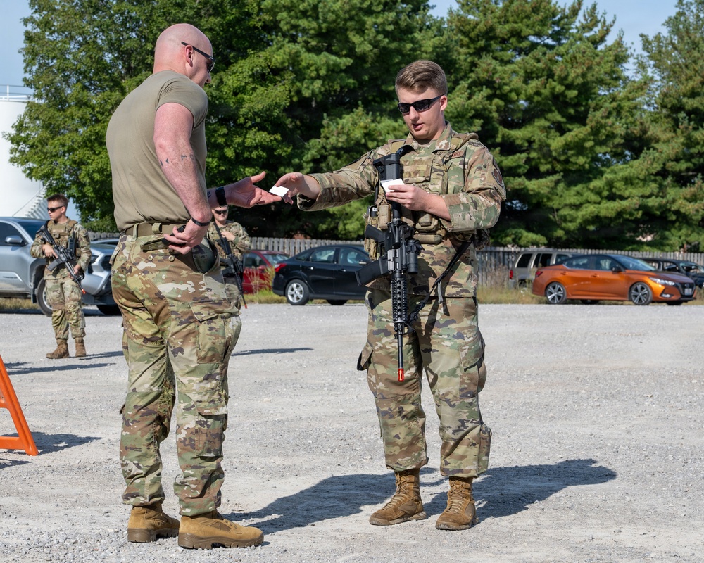 Two military personnel in OCP (Operational Camouflage Pattern) uniforms are engaged in what appears to be an identification or credential check at what looks like a security checkpoint in a gravel parking area. The armed soldier on the right is holding a rifle and exchanging documents or cards with the bald soldier on the left. Additional armed personnel are visible in the background near parked civilian vehicles.