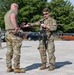Two military personnel in OCP (Operational Camouflage Pattern) uniforms are engaged in what appears to be an identification or credential check at what looks like a security checkpoint in a gravel parking area. The armed soldier on the right is holding a rifle and exchanging documents or cards with the bald soldier on the left. Additional armed personnel are visible in the background near parked civilian vehicles.