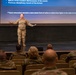 A female military service member in OCP (Operational Camouflage Pattern) uniform is delivering a presentation on a stage to an audience of uniformed personnel seated in rows. The presentation slide behind her discusses disciplinary procedures, referencing 'Airman' which indicates a U.S. Air Force context. The setting appears to be a military auditorium or theater used for training or briefing purposes.
