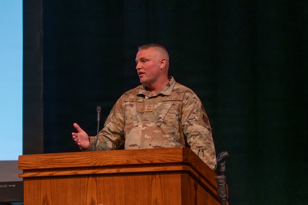 A U.S. Air Force service member in OCP (Operational Camouflage Pattern) uniform is speaking at a wooden podium, likely during a formal briefing or ceremony. The individual appears to be a senior non-commissioned officer based on visible rank insignia on the chest. The setting appears to be an auditorium or conference hall with a dark curtain backdrop and a projection screen visible to the left.