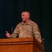 A U.S. Air Force service member in OCP (Operational Camouflage Pattern) uniform is speaking at a wooden podium, likely during a formal briefing or ceremony. The individual appears to be a senior non-commissioned officer based on visible rank insignia on the chest. The setting appears to be an auditorium or conference hall with a dark curtain backdrop and a projection screen visible to the left.