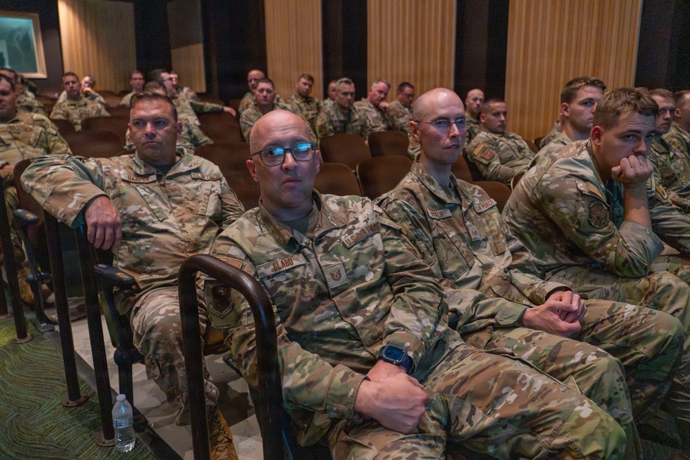 A large group of U.S. Air Force personnel in OCP (Operational Camouflage Pattern) uniforms are seated in an auditorium or briefing room, attentively watching a presentation or briefing. The foreground shows several airmen clearly, including one wearing blue-framed glasses with a name tape reading 'GLADU' and another reading 'HARPER'. The setting appears to be a formal military gathering or training event.