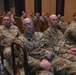 A large group of U.S. Air Force personnel in OCP (Operational Camouflage Pattern) uniforms are seated in an auditorium or briefing room, attentively watching a presentation or briefing. The foreground shows several airmen clearly, including one wearing blue-framed glasses with a name tape reading 'GLADU' and another reading 'HARPER'. The setting appears to be a formal military gathering or training event.