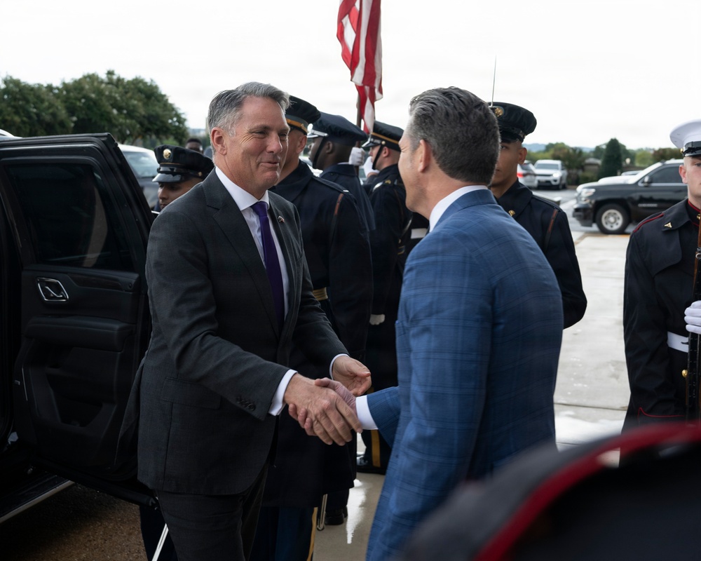 Two men in civilian business attire are shaking hands in what appears to be an official arrival ceremony. A military honor guard in dress uniforms is lined up in the background, and a U.S. flag is prominently displayed. The setting suggests a formal government or military installation welcome event.