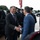 Two men in civilian business attire are shaking hands in what appears to be an official arrival ceremony. A military honor guard in dress uniforms is lined up in the background, and a U.S. flag is prominently displayed. The setting suggests a formal government or military installation welcome event.