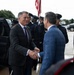 Two men in civilian business attire are shaking hands in what appears to be an official arrival ceremony. A military honor guard in dress uniforms is lined up in the background, and a U.S. flag is prominently displayed. The setting suggests a formal government or military installation welcome event.