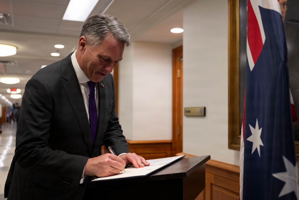 A man in a dark suit and purple tie is signing a document at a lectern or writing stand. An Australian flag is prominently visible to his right, suggesting an official or diplomatic setting. The scene appears to take place in a government or institutional building corridor.