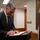 A man in a dark suit and purple tie is signing a document at a lectern or writing stand. An Australian flag is prominently visible to his right, suggesting an official or diplomatic setting. The scene appears to take place in a government or institutional building corridor.