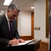 A man in a dark suit and purple tie is signing a document at a lectern or writing stand. An Australian flag is prominently visible to his right, suggesting an official or diplomatic setting. The scene appears to take place in a government or institutional building corridor.