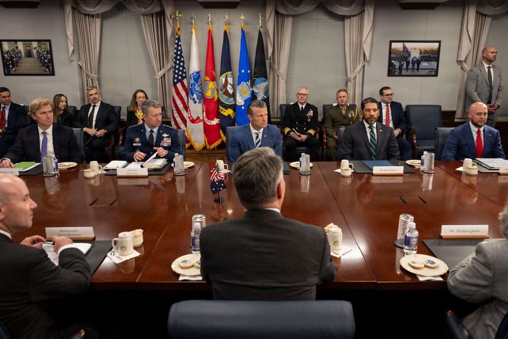 A formal high-level government or military meeting is taking place around a large conference table, likely at the Pentagon or a similar U.S. defense facility. Multiple officials in both civilian attire and military uniforms are seated, with name placards visible on the table. Several U.S. military branch flags and the American flag are displayed prominently in the background.