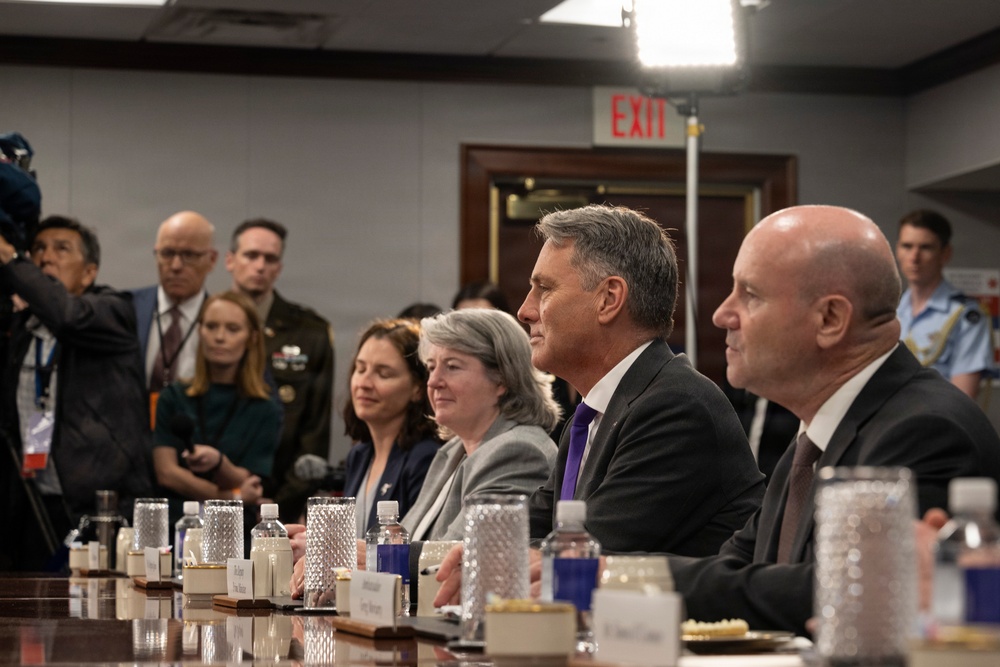 A formal meeting or briefing is taking place in what appears to be a government or official conference room. Several individuals are seated at a long conference table with water bottles, glasses, and name placards visible. Media personnel with cameras are present in the background, suggesting this is an official or press-accessible event.