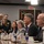 A formal meeting or briefing is taking place in what appears to be a government or official conference room. Several individuals are seated at a long conference table with water bottles, glasses, and name placards visible. Media personnel with cameras are present in the background, suggesting this is an official or press-accessible event.