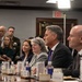 A formal meeting or briefing is taking place in what appears to be a government or official conference room. Several individuals are seated at a long conference table with water bottles, glasses, and name placards visible. Media personnel with cameras are present in the background, suggesting this is an official or press-accessible event.