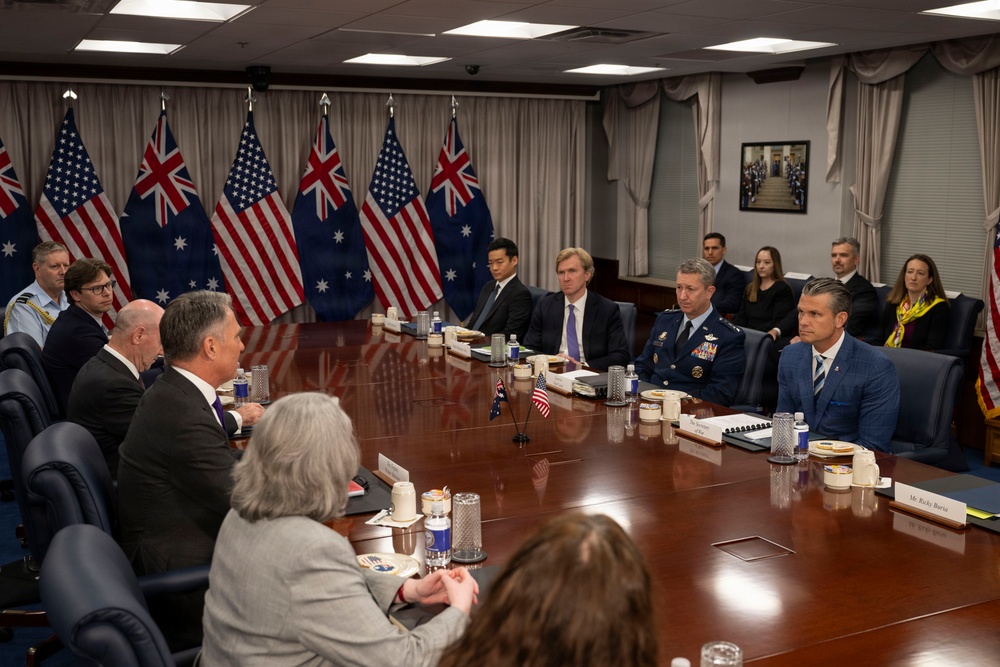 A formal bilateral meeting between United States and Australian officials is taking place in what appears to be a government conference room, likely the Pentagon or a similar high-level facility. Participants are seated around a large oval conference table with both American and Australian flags displayed prominently in the background. The setting suggests a high-level diplomatic or defense meeting between the two allied nations.