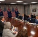 A formal bilateral meeting between United States and Australian officials is taking place in what appears to be a government conference room, likely the Pentagon or a similar high-level facility. Participants are seated around a large oval conference table with both American and Australian flags displayed prominently in the background. The setting suggests a high-level diplomatic or defense meeting between the two allied nations.