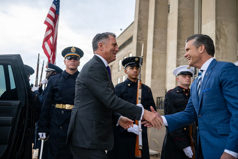 Two men in civilian business attire are shaking hands in what appears to be an official ceremonial greeting outside a large government-style building. Military honor guard personnel from multiple branches are standing at attention in the background, holding ceremonial rifles. An American flag is visible, suggesting a formal diplomatic or official reception event.