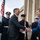 Two men in civilian business attire are shaking hands in what appears to be an official ceremonial greeting outside a large government-style building. Military honor guard personnel from multiple branches are standing at attention in the background, holding ceremonial rifles. An American flag is visible, suggesting a formal diplomatic or official reception event.