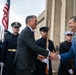 Two men in civilian business attire are shaking hands in what appears to be an official ceremonial greeting outside a large government-style building. Military honor guard personnel from multiple branches are standing at attention in the background, holding ceremonial rifles. An American flag is visible, suggesting a formal diplomatic or official reception event.