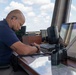 A bald man wearing a navy blue shirt and a headset is seated at a workstation in what appears to be an air traffic control tower. He is focused on writing or reviewing documents while surrounded by multiple monitors, a keyboard, binoculars, and communication equipment. Large windows behind him reveal a partly cloudy sky, consistent with an elevated control tower environment.