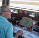 An older male air traffic controller is seated at a control tower workstation, monitoring multiple screens and equipment. The workspace contains radar displays, digital clocks showing local time 13:30 and Zulu time 18:30, binoculars, and various control terminals. A runway or taxiway is visible through the large windows behind him.