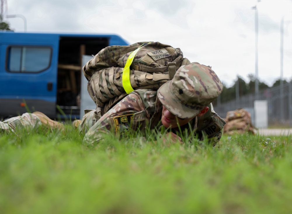 A U.S. Army Military Police soldier in OCP (Operational Camouflage Pattern) uniform is prone on grass, appearing to be engaged in a ground combat training exercise or detainee control drill. The soldier is wearing full gear including a tactical vest with a high-visibility yellow strap, and has 'MP' visible on their uniform. The setting appears to be a military installation or training area in a European environment.