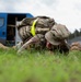 A U.S. Army Military Police soldier in OCP (Operational Camouflage Pattern) uniform is prone on grass, appearing to be engaged in a ground combat training exercise or detainee control drill. The soldier is wearing full gear including a tactical vest with a high-visibility yellow strap, and has 'MP' visible on their uniform. The setting appears to be a military installation or training area in a European environment.
