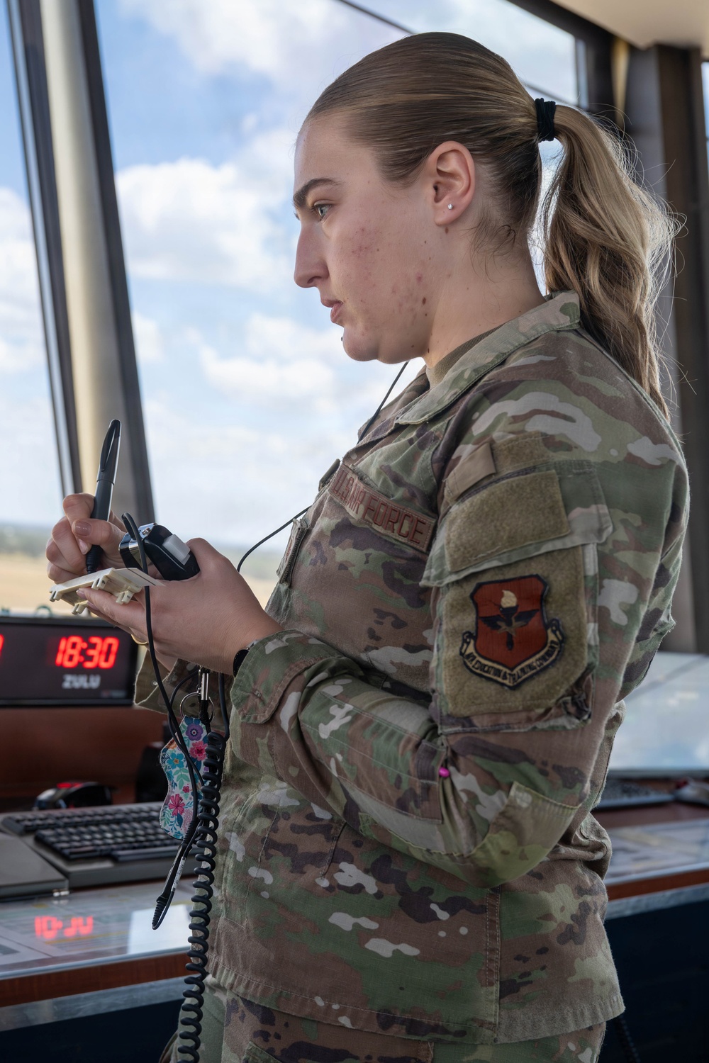 A female U.S. Air Force service member in Operational Camouflage Pattern (OCP) uniform is standing in what appears to be an air traffic control tower, holding a radio/communication handset. She is wearing a unit patch on her left sleeve and a name tape reading 'AIR FORCE' on her chest. A digital clock in the background displays 18:30 Zulu time.