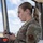A female U.S. Air Force service member in Operational Camouflage Pattern (OCP) uniform is standing in what appears to be an air traffic control tower, holding a radio/communication handset. She is wearing a unit patch on her left sleeve and a name tape reading 'AIR FORCE' on her chest. A digital clock in the background displays 18:30 Zulu time.