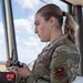 A female U.S. Air Force service member in Operational Camouflage Pattern (OCP) uniform is standing in what appears to be an air traffic control tower, holding a radio/communication handset. She is wearing a unit patch on her left sleeve and a name tape reading 'AIR FORCE' on her chest. A digital clock in the background displays 18:30 Zulu time.