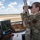 A female U.S. Air Force airman in OCP (Operational Camouflage Pattern) uniform is using binoculars to observe the airfield from what appears to be an air traffic control tower. She is stationed at a workstation equipped with radar displays, keyboards, and communication equipment, with a clear view of the runway through large windows. The setting is consistent with active air traffic control operations at a military airbase.